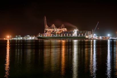 A large cargo ship is docked at a bustling port during nighttime, with cranes towering over it. The scene is illuminated by artificial lights, casting reflections on the calm water. Containers are stacked high on the ship, indicating active maritime trade.