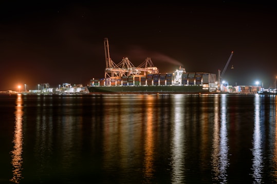 A large cargo ship is docked at a bustling port during nighttime, with cranes towering over it. The scene is illuminated by artificial lights, casting reflections on the calm water. Containers are stacked high on the ship, indicating active maritime trade.