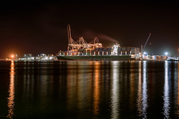 A large cargo ship is docked at a bustling port during nighttime, with cranes towering over it. The scene is illuminated by artificial lights, casting reflections on the calm water. Containers are stacked high on the ship, indicating active maritime trade.