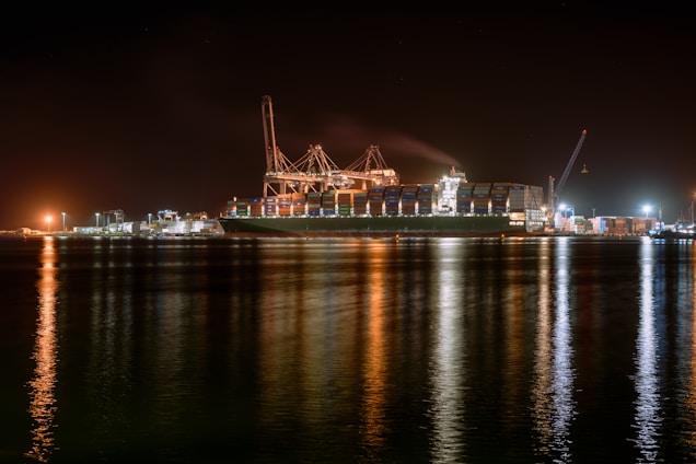 A large cargo ship is docked at a bustling port during nighttime, with cranes towering over it. The scene is illuminated by artificial lights, casting reflections on the calm water. Containers are stacked high on the ship, indicating active maritime trade.
