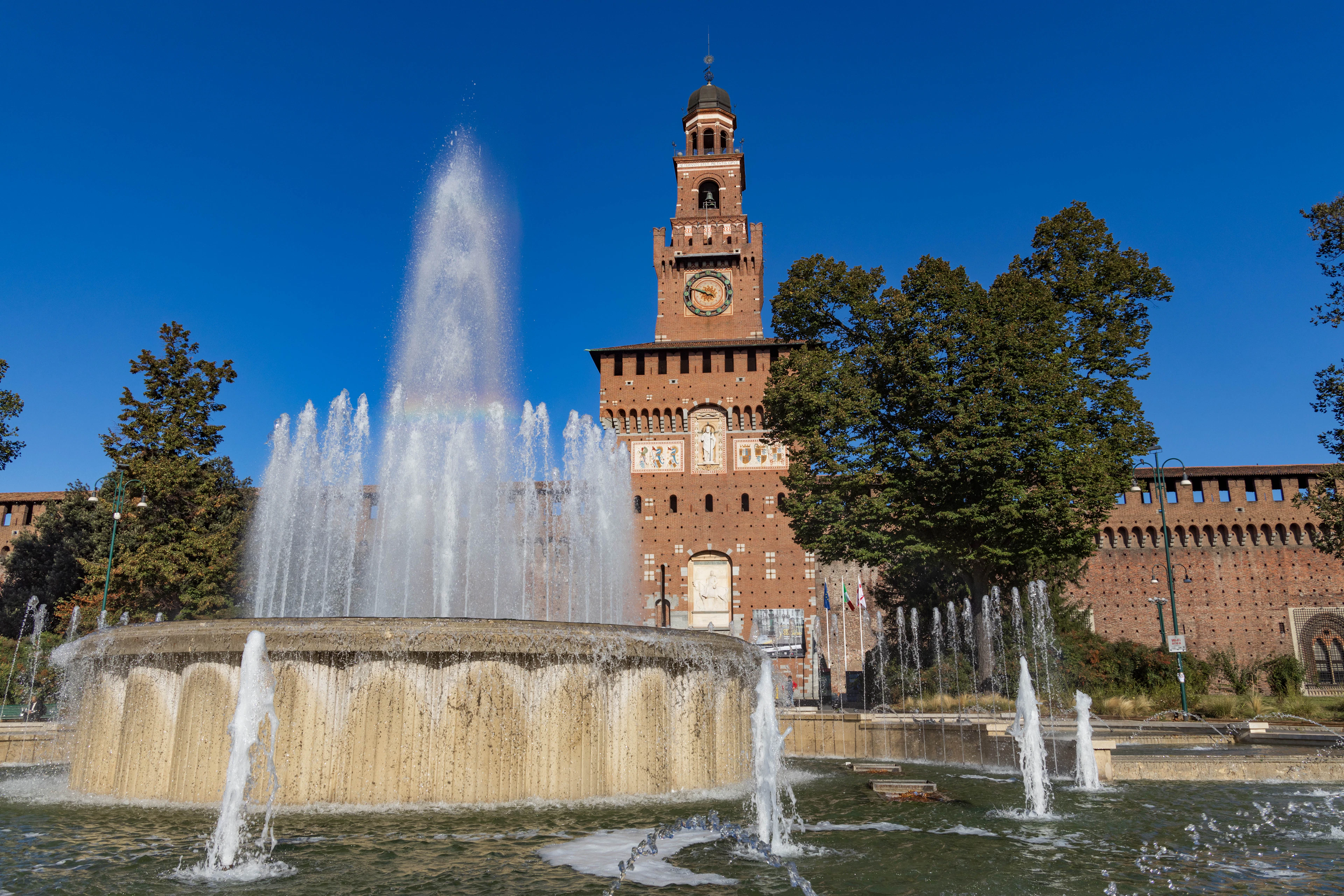 The red-brick Filarete Tower of Castello Sforzesco rising above the jets of the fountain on Piazza Castello in Milan, framed by summer trees under a deep blue sky.