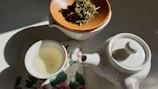 A white ceramic teapot and a teacup with light-colored tea sit on a floral-patterned cloth. Next to them is a wooden dish containing loose leaf tea, positioned in soft natural lighting.