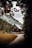 A picturesque wooden chalet is nestled among tall, dense coniferous trees with a mountain cliff in the background. The scene is framed by autumnal leaves at the top, adding a sense of enclosure. A winding gravel path leads towards the house, inviting a sense of journey or arrival.