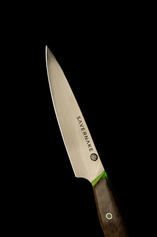 A sleek, sharpened knife with a polished metallic blade and a dark wooden handle, set against a stark black background. The knife has the brand name Savernake inscribed on it, and there is a green accent between the blade and handle.