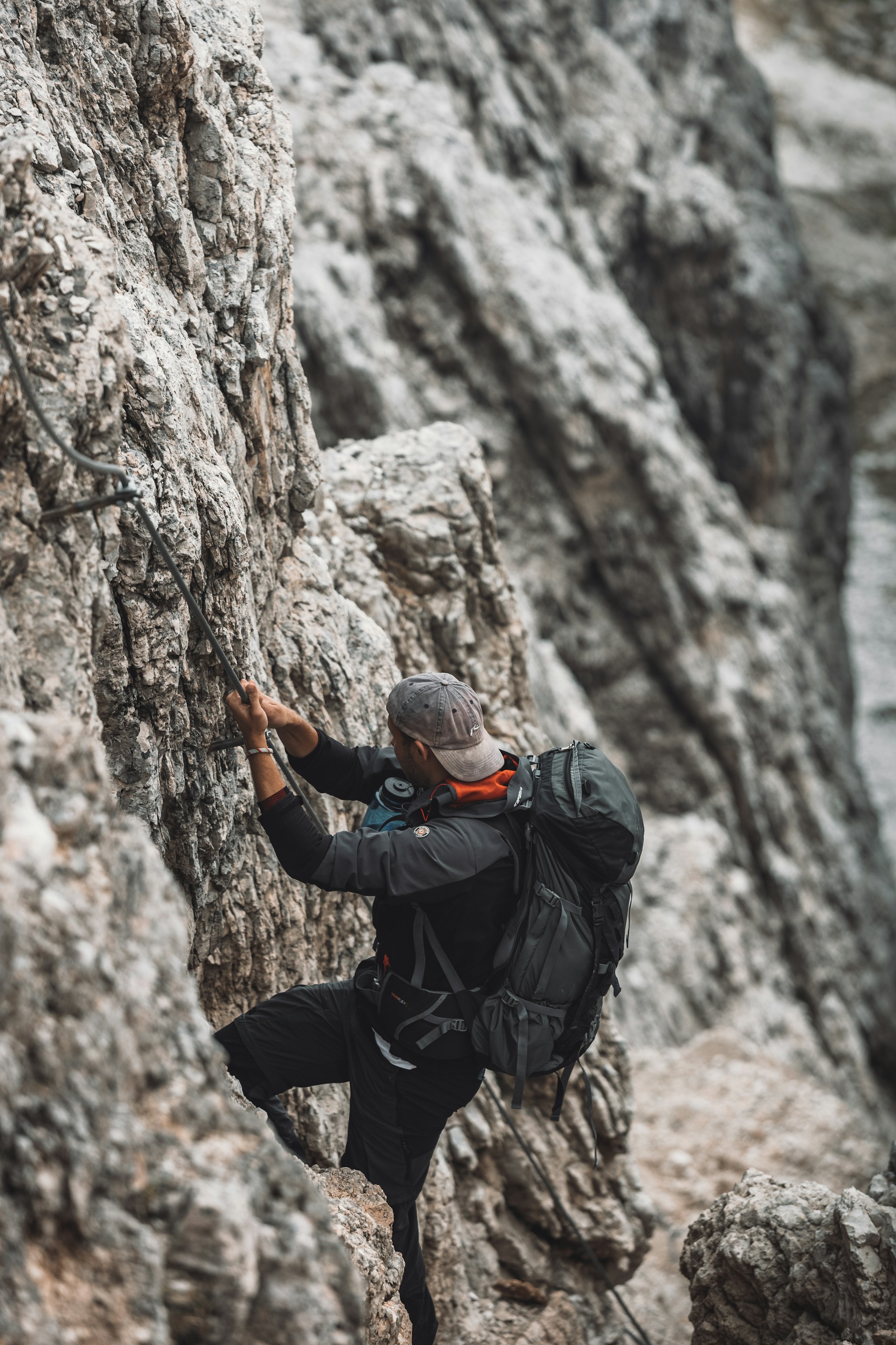 A person wearing outdoor climbing gear is scaling a rugged rocky surface, using a metal cable for support. The individual is dressed in dark clothing with a backpack and is focused on ascending the mountainous terrain.