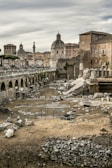 Ancient Roman ruins with scattered stone blocks and remnants of columns fill the foreground. In the background, historic buildings with domed roofs and a tall column create a classical cityscape under a cloudy sky.