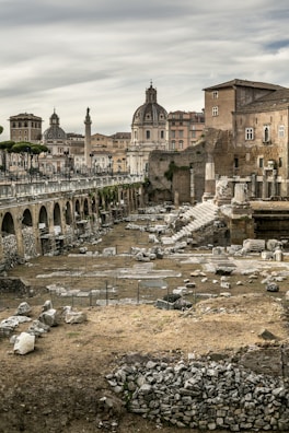 Ancient Roman ruins with scattered stone blocks and remnants of columns fill the foreground. In the background, historic buildings with domed roofs and a tall column create a classical cityscape under a cloudy sky.