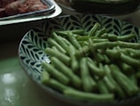 A plate filled with fresh green beans in a bowl with a blue and white leaf pattern. In the background, there are trays containing raw meat.