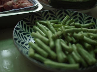 A plate filled with fresh green beans in a bowl with a blue and white leaf pattern. In the background, there are trays containing raw meat.