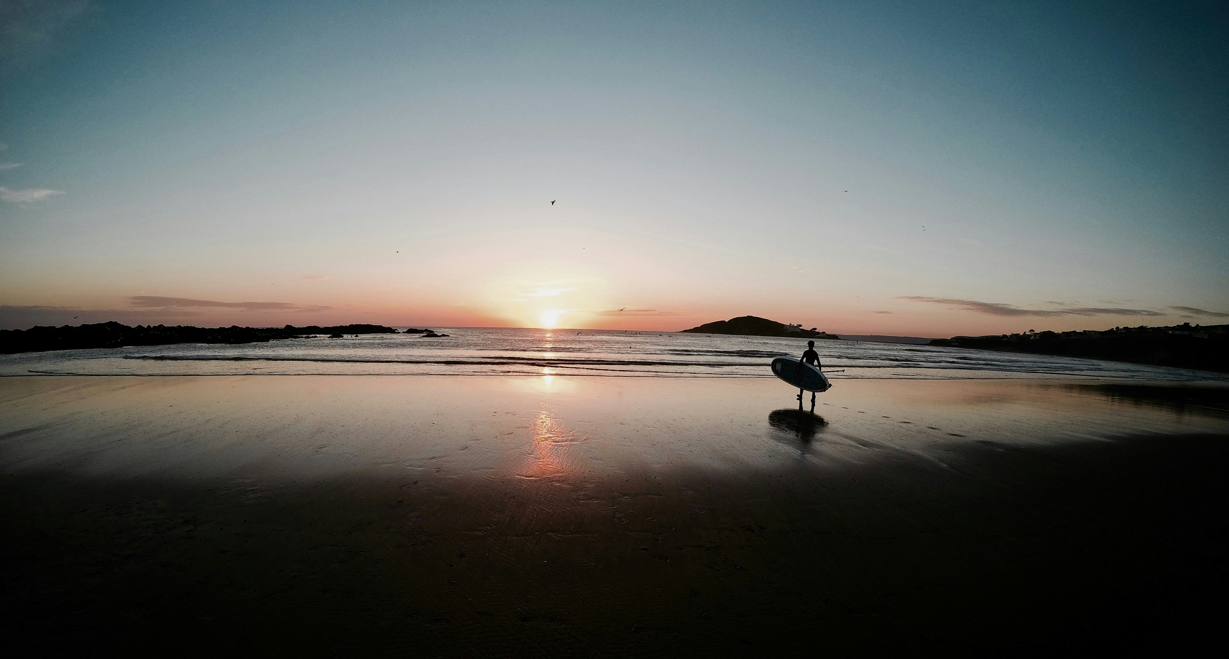 silhouette of person carrying surfboard walking on beach during sunset, solo surf @ sunset