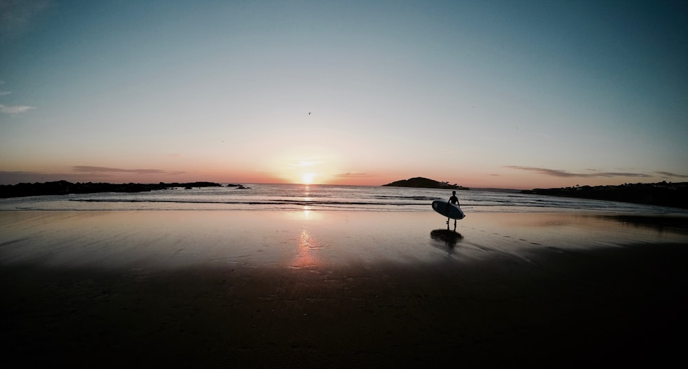 silhouette of person carrying surfboard walking on beach during sunset