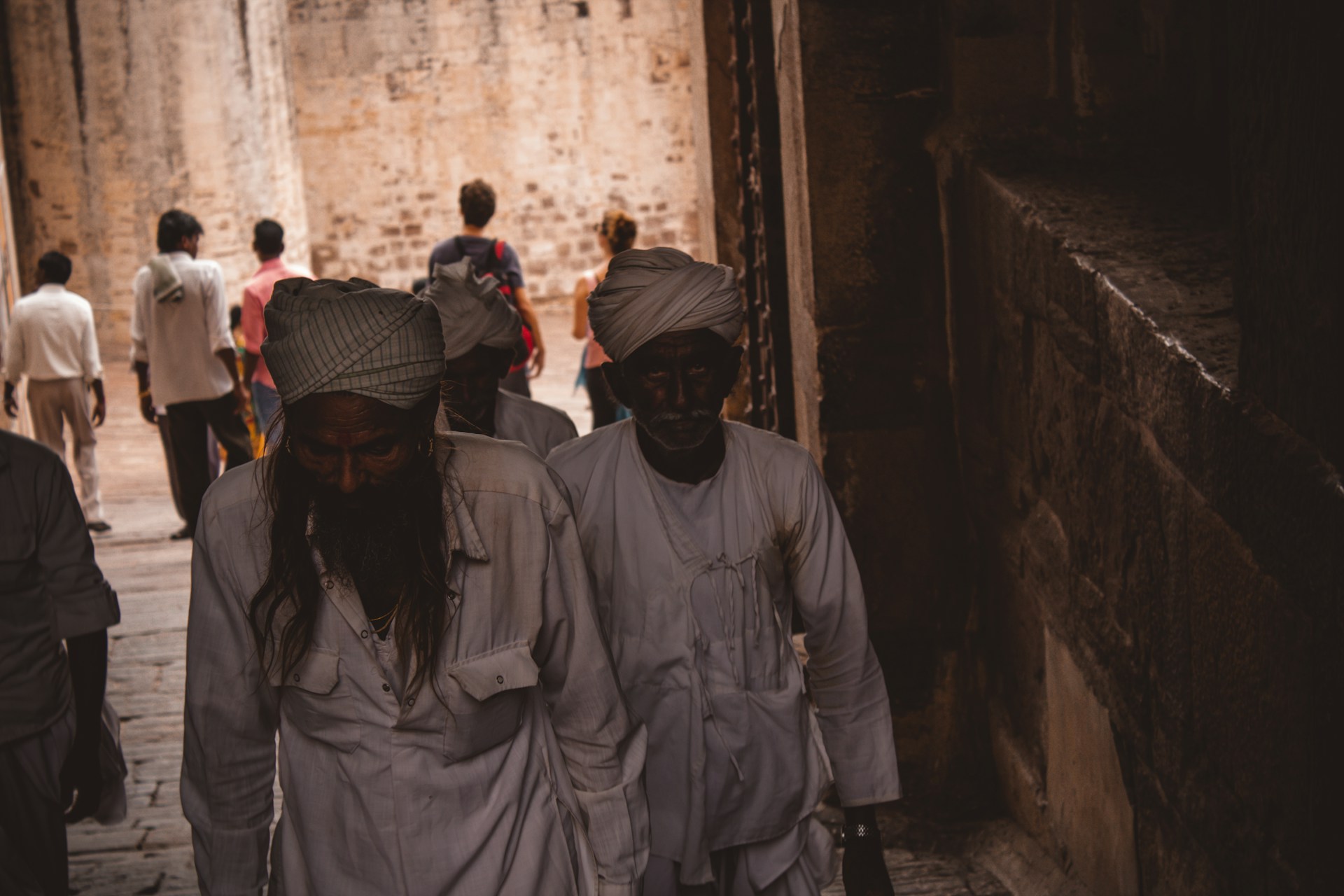 Two elderly men wearing traditional white garments and turbans walk through an ancient stone corridor. Several other people are visible in the background, adding life to the rustic setting. The dim lighting casts shadows, enhancing the texture of the stone walls.