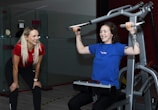 Two women are in a gym setting, one is standing and smiling, wearing a red shirt and black pants, likely a trainer. The other is seated on a workout machine, using it while wearing a blue shirt, and smiling. The background includes a glass partition and gym equipment.