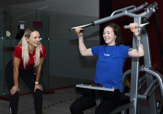Two women are in a gym setting, one is standing and smiling, wearing a red shirt and black pants, likely a trainer. The other is seated on a workout machine, using it while wearing a blue shirt, and smiling. The background includes a glass partition and gym equipment.