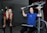 Two women are in a gym setting, one is standing and smiling, wearing a red shirt and black pants, likely a trainer. The other is seated on a workout machine, using it while wearing a blue shirt, and smiling. The background includes a glass partition and gym equipment.