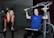 Two women are in a gym setting, one is standing and smiling, wearing a red shirt and black pants, likely a trainer. The other is seated on a workout machine, using it while wearing a blue shirt, and smiling. The background includes a glass partition and gym equipment.