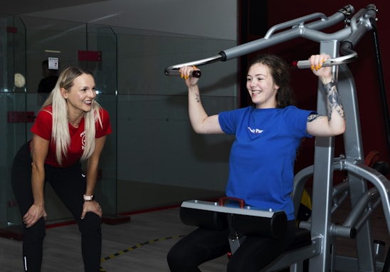 Two women are in a gym setting, one is standing and smiling, wearing a red shirt and black pants, likely a trainer. The other is seated on a workout machine, using it while wearing a blue shirt, and smiling. The background includes a glass partition and gym equipment.