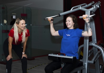 Two women are in a gym setting, one is standing and smiling, wearing a red shirt and black pants, likely a trainer. The other is seated on a workout machine, using it while wearing a blue shirt, and smiling. The background includes a glass partition and gym equipment.