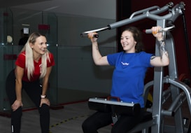 Two women are in a gym setting, one is standing and smiling, wearing a red shirt and black pants, likely a trainer. The other is seated on a workout machine, using it while wearing a blue shirt, and smiling. The background includes a glass partition and gym equipment.