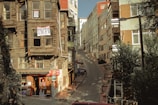 A narrow, steep street flanked by aging wooden houses with signs indicating they are for sale. On the left, a small convenience store is situated at the base of one of the houses, displaying various goods. Modern multi-story buildings line the right side of the street. Shadows and sunlight create an interplay of light and dark across the scene.