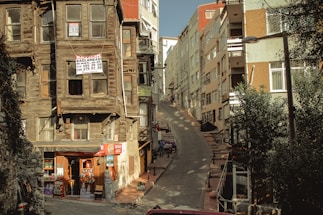 A narrow, steep street flanked by aging wooden houses with signs indicating they are for sale. On the left, a small convenience store is situated at the base of one of the houses, displaying various goods. Modern multi-story buildings line the right side of the street. Shadows and sunlight create an interplay of light and dark across the scene.