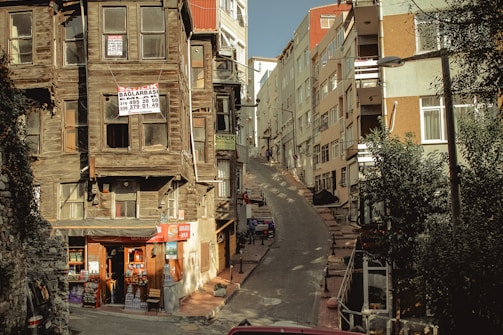 A narrow, steep street flanked by aging wooden houses with signs indicating they are for sale. On the left, a small convenience store is situated at the base of one of the houses, displaying various goods. Modern multi-story buildings line the right side of the street. Shadows and sunlight create an interplay of light and dark across the scene.