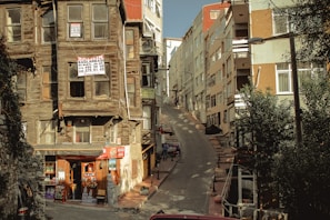 A narrow, steep street flanked by aging wooden houses with signs indicating they are for sale. On the left, a small convenience store is situated at the base of one of the houses, displaying various goods. Modern multi-story buildings line the right side of the street. Shadows and sunlight create an interplay of light and dark across the scene.