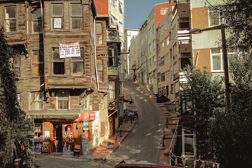 A narrow, steep street flanked by aging wooden houses with signs indicating they are for sale. On the left, a small convenience store is situated at the base of one of the houses, displaying various goods. Modern multi-story buildings line the right side of the street. Shadows and sunlight create an interplay of light and dark across the scene.