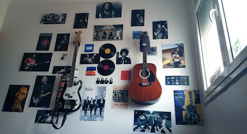 A wall decorated with numerous posters and pictures of musicians and bands, such as The Beatles. Several guitars hang on the wall amidst the artwork, including an acoustic and an electric guitar. There are vinyl records displayed as well, adding to the musical theme. A window is partially visible on the right, allowing natural light into the space, which enhances the artistic display.