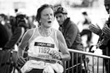A black and white image of a marathon runner with a focused and determined expression, wearing a sleeveless jersey labeled with race number 1162 and a name tag. The runner is being cheered on by spectators in the background, who are clapping.