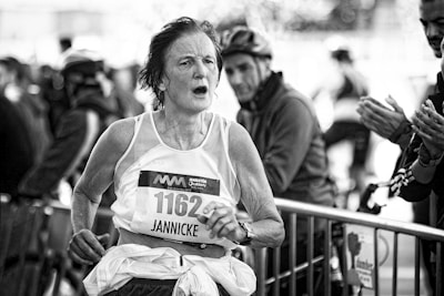 A black and white image of a marathon runner with a focused and determined expression, wearing a sleeveless jersey labeled with race number 1162 and a name tag. The runner is being cheered on by spectators in the background, who are clapping.