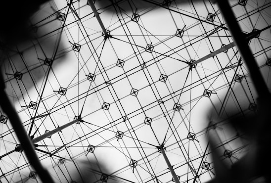 A complex network of interconnected wires and nodes forms a geometric grid pattern against a bright background. The structure appears intricate and symmetrical, with intersecting lines creating diamond shapes.