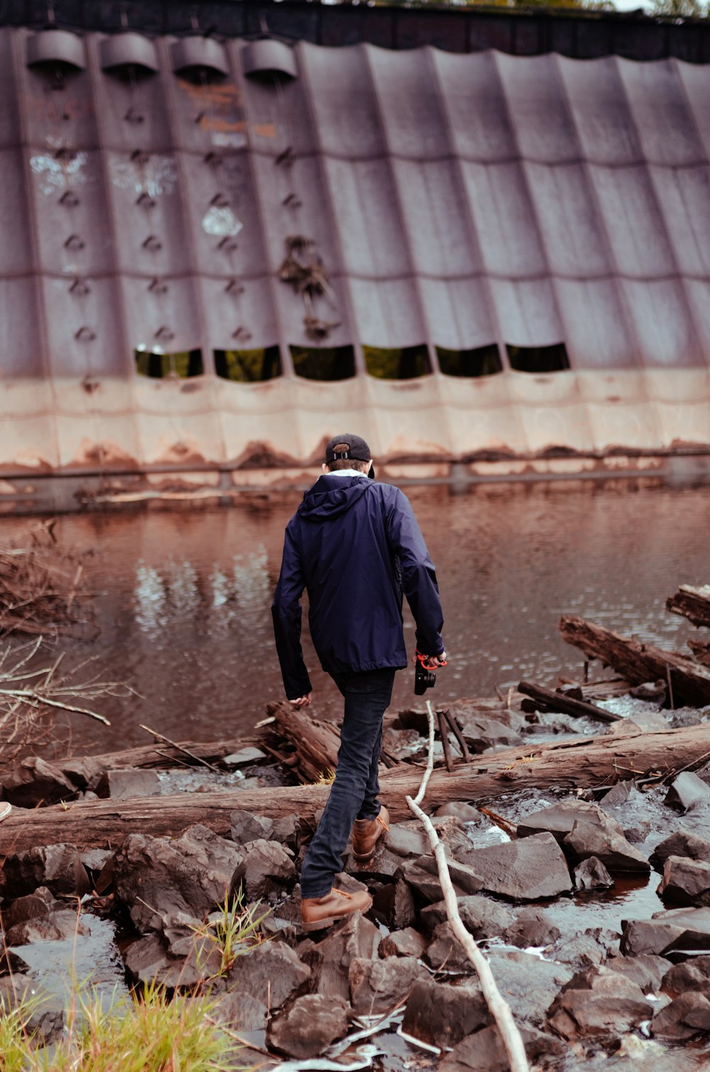 man in blue jacket and black pants standing on brown wood log near body of water