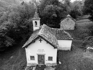 A small, historic chapel with a stone-tiled roof and a modest bell tower is situated in a dense forest clearing. The structure features a wooden door, above which a cross and a Latin inscription are visible. The surrounding vegetation is lush and envelops the chapel, adding a serene and isolated atmosphere.