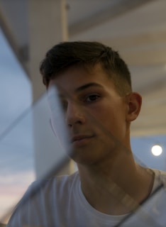 A young person is partially visible through a glass surface, which reflects some light. The setting appears to be outdoors or near a large window, with a soft and diffused sky visible in the background. The lighting is dim, contributing to a contemplative atmosphere.