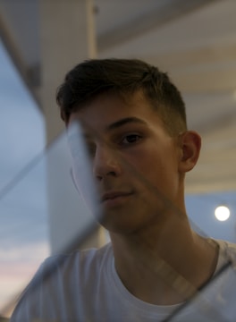 A young person is partially visible through a glass surface, which reflects some light. The setting appears to be outdoors or near a large window, with a soft and diffused sky visible in the background. The lighting is dim, contributing to a contemplative atmosphere.