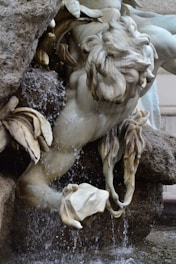 A detailed stone sculpture of a figure partially submerged in water, with intricate carvings of flowing hair and textured surfaces. Water cascades around the statue, enhancing the dynamic pose of the figure, which appears to be part of a larger fountain structure. The sculpture showcases skillful craftsmanship with a focus on movement and fluidity.