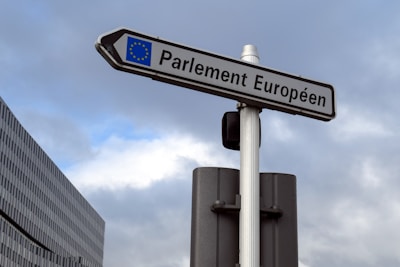 A street sign with the text 'Parlement Européen' is mounted on a pole against a backdrop of a cloudy sky. The sign also features the European Union flag. A modern building with a grid-like facade is partially visible to the left.