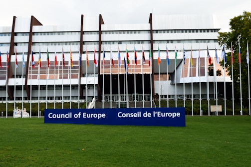 A modern architectural building with a series of vertical and horizontal elements creates a striking facade. Numerous international flags are displayed on flagpoles at the front, and a large blue sign with the words 'Council of Europe' and 'Conseil de l'Europe' is prominent in the foreground. The building is surrounded by a well-maintained green lawn.