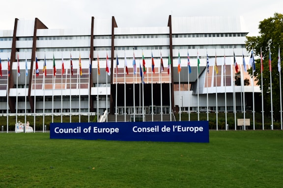 A modern architectural building with a series of vertical and horizontal elements creates a striking facade. Numerous international flags are displayed on flagpoles at the front, and a large blue sign with the words 'Council of Europe' and 'Conseil de l'Europe' is prominent in the foreground. The building is surrounded by a well-maintained green lawn.