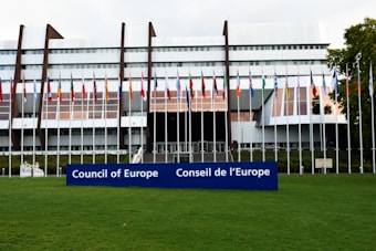 A modern architectural building with a series of vertical and horizontal elements creates a striking facade. Numerous international flags are displayed on flagpoles at the front, and a large blue sign with the words 'Council of Europe' and 'Conseil de l'Europe' is prominent in the foreground. The building is surrounded by a well-maintained green lawn.