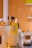 A stack of bright yellow paper cups with Chinese characters printed on them, placed on a counter. There are black utensils and a ceramic container with a similar design theme present. The background consists of an orange tiled wall with shelves showing small items.