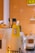 A stack of bright yellow paper cups with Chinese characters printed on them, placed on a counter. There are black utensils and a ceramic container with a similar design theme present. The background consists of an orange tiled wall with shelves showing small items.