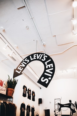 An interior retail space featuring a large Levi's sign hanging from the ceiling. The room is well-lit with a modern design, featuring white walls and ceiling. On the wall to the left, several black T-shirts are displayed, each having a colorful graphic print. There is a small potted plant on a shelf below the T-shirts. To the right, an old-fashioned Singer sewing machine is part of the decor.