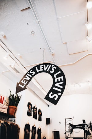 An interior retail space featuring a large Levi's sign hanging from the ceiling. The room is well-lit with a modern design, featuring white walls and ceiling. On the wall to the left, several black T-shirts are displayed, each having a colorful graphic print. There is a small potted plant on a shelf below the T-shirts. To the right, an old-fashioned Singer sewing machine is part of the decor.