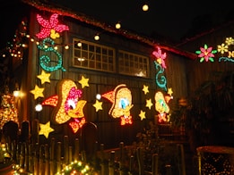 A festive display with cartoon characters and stars illuminated with colorful lights. The characters are adorned with Santa hats, and there are decorative light patterns and stars surrounding them. The scene is set on the exterior of a wooden building, with additional festive lighting and garlands.