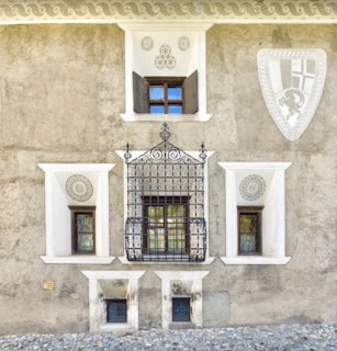 A textured stone wall with three windows of varying sizes, each having ornate wrought iron grills. The wall is adorned with decorative geometric carvings and a large shield-like emblem on the right side. The base of the wall is lined with cobblestones, adding to the rustic architectural aesthetic.