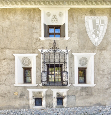 A textured stone wall with three windows of varying sizes, each having ornate wrought iron grills. The wall is adorned with decorative geometric carvings and a large shield-like emblem on the right side. The base of the wall is lined with cobblestones, adding to the rustic architectural aesthetic.