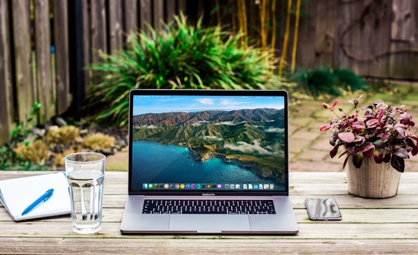 A workspace is set on a wooden table in an outdoor garden setting. A laptop displaying a scenic mountain and ocean wallpaper is centrally placed. To the left is a glass of water and an open notebook with a blue pen. On the right side, a smartphone lies next to a small potted plant with red-tinted leaves. The background features a garden with green plants and a wooden fence.