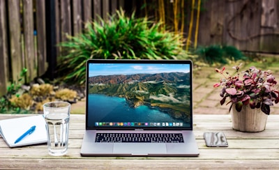 A workspace is set on a wooden table in an outdoor garden setting. A laptop displaying a scenic mountain and ocean wallpaper is centrally placed. To the left is a glass of water and an open notebook with a blue pen. On the right side, a smartphone lies next to a small potted plant with red-tinted leaves. The background features a garden with green plants and a wooden fence.